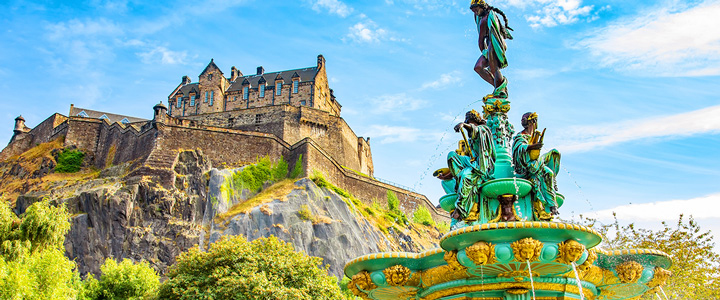 Edinburgh Castle thront über den Princes Street Gardens. Im Vordergrund steht die kunstvoll gestaltete Ross Fountain, umgeben von grünen Rasenflächen.